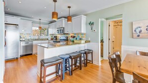 A modern kitchen with white cabinets, a central island with stools, and a dining area with a wooden table and chairs. - Oceanfront, event-friendly home with multiple decks & WiFi - 2 dogs ok (Folly Beach)