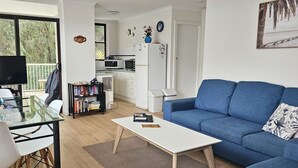 A compact living space with a sofa, a coffee table, a bookshelf, a kitchen area, and a balcony with a view of trees.