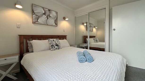 A neatly made bed with a patterned bedspread, two rolled towels, and a wall-mounted artwork.