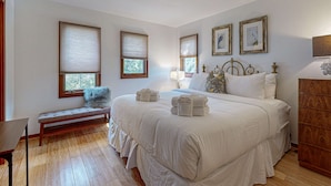 Un dormitorio con una cama grande, un banco, un aparador de madera y dos ventanas con persianas.
