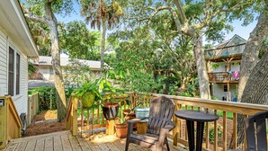 A wooden deck with a chair and table, surrounded by potted plants and trees. - One Block to the Beach Cute Home w/ Deck, Garden Patio, Central AC & WiFi (Folly Beach)