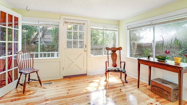 A room with wooden flooring, a white door, and large windows with a view of greenery outside. - One Block to the Beach Cute Home w/ Deck, Garden Patio, Central AC & WiFi (Folly Beach)