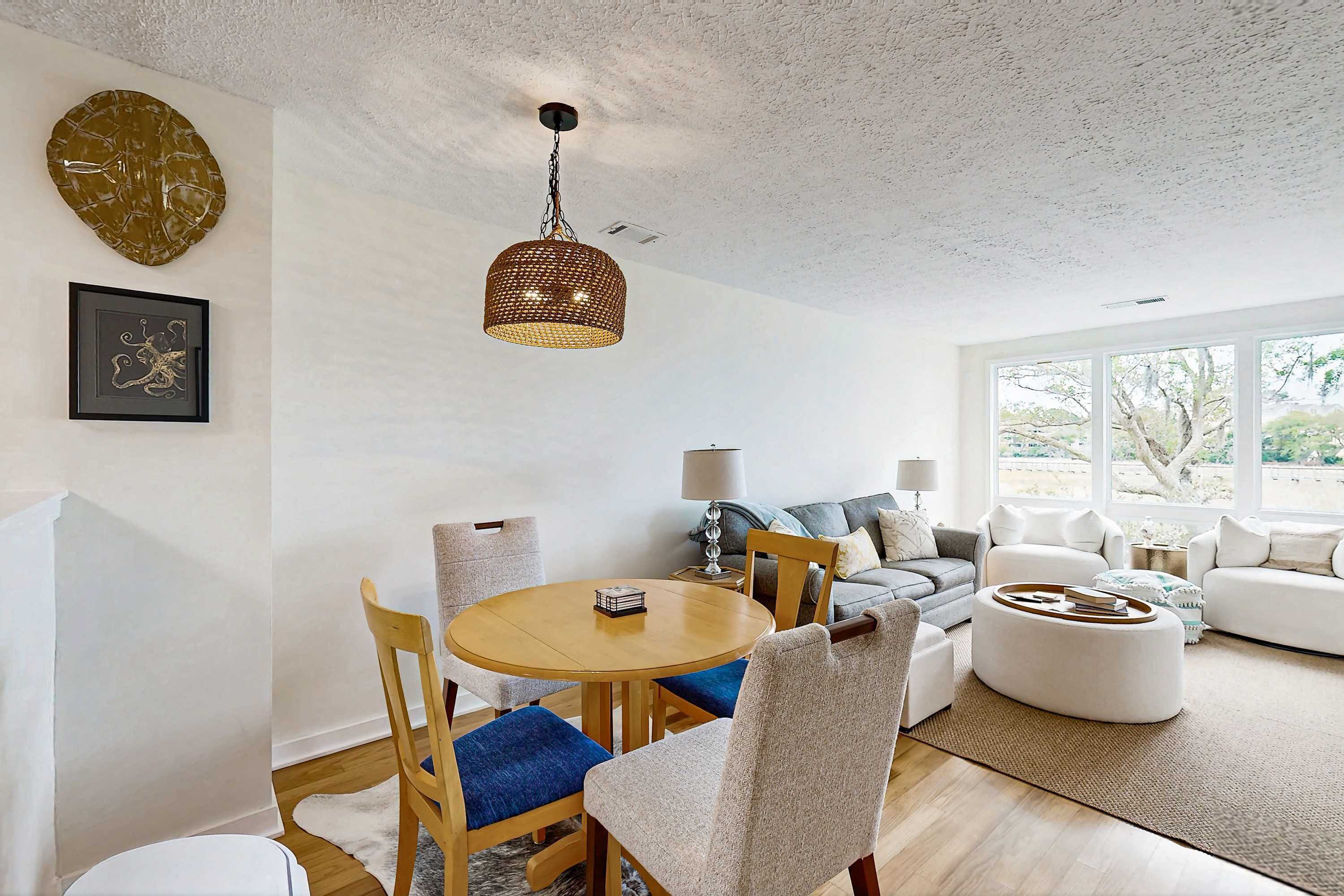 A modern living room with a round wooden table, blue cushioned chairs, a white sofa, and a white armchair. There is a large window providing natural light and a view of trees.