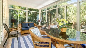 Ein Wintergarten mit Korbmöbeln, einem Glastisch und großen Fenstern mit Blick auf üppiges tropisches Grün.