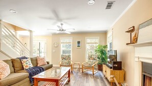 Ein geräumiges Wohnzimmer mit Holzboden, einem Glastisch, einer Couch mit bunten Kissen, einem Kamin und einem Deckenventilator.
