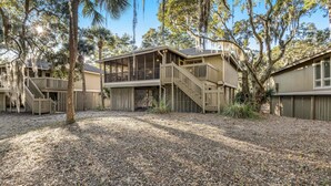 A wooden cabin with a balcony and a covered porch, surrounded by trees and a gravel ground. - Charming Island Getaway with a Furnished Deck Offering Forest & Fairway Views (Seabrook Island)