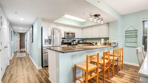 A modern kitchen with white cabinets, a central island with bar stools, stainless steel appliances, and a ceiling light fixture. - Updated oceanfront condo w/shared pools & views - just steps from the beach (Folly Beach)