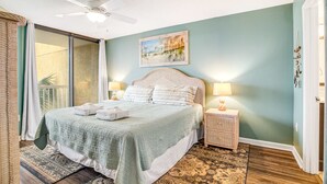A bedroom with a bed, two bedside tables, a ceiling fan, a painting, and a view through a sliding door. - Updated oceanfront condo w/shared pools & views - just steps from the beach (Folly Beach)
