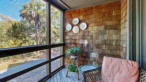 Eine Holzterrasse mit einem Korbstuhl, einem pinken Kissen, einem Blumentopf und einer Wand mit dekorativen Tellern.
