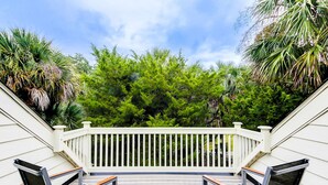 Una terraza de madera con dos sillas de metal y una barandilla blanca, rodeada de frondosos árboles verdes.