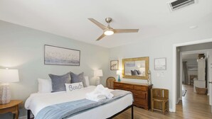 Una habitación de hotel con cama, ventilador de techo, mesita de noche con lámpara, cómoda y espejo.
