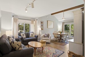 Ein geräumiges Wohnzimmer mit einer Couch, Sesseln, einem Couchtisch und einem Essbereich mit Tisch und Stühlen. Große Fenster sorgen für natürliches Licht und Ausblick ins Freie.