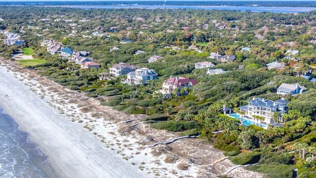 An aerial view of a coastal residential area with houses, a beach, and a large body of water in the background.