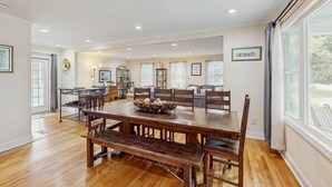 A spacious dining area with a large wooden table and benches, a kitchen in the background, and large windows with curtains.