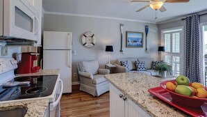 A kitchen with a microwave, stove, and red coffee maker. There is a chair and a table with a lamp. A clock and a framed picture are on the wall. - Beachfront condo with pool, beach access, pier, private balcony, & central AC (Isle of Palms)