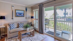 A living room with a sofa, a coffee table, a lamp, a ceiling fan, a framed picture, and a view of the outdoors through a sliding glass door. - Beachfront condo with pool, beach access, pier, private balcony, & central AC (Isle of Palms)