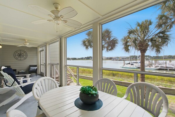 A covered outdoor seating area with a table and chairs, a ceiling fan, and a view of a lake with boats and palm trees.