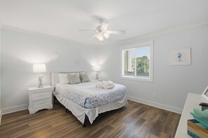 Een slaapkamer met een bed, nachtkastjes, een plafondventilator, een raam met jaloezieën en een wandklok.