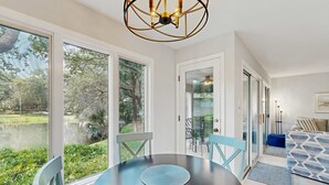 Un comedor con una mesa redonda y sillas, ventanas amplias con vistas a zonas verdes y un estanque, y una lámpara de araña moderna.