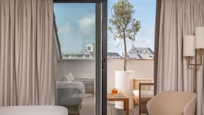 Una habitación de hotel con una cama, una silla, una mesa con lámpara y vistas a la ciudad a través de un ventanal.