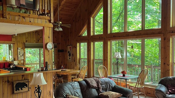 A cozy cabin interior with a wooden ceiling, large windows, a dining area, and a living room with a leather sofa and ottoman. - Hartford Hideaway! Soothing cabin in the Smokies. (Hartford)