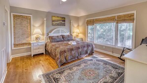 A bedroom with a large bed, two bedside tables with lamps, a ceiling fan, and a view of trees through the windows. - Remodeled Home with Deck, Sun Porch & In-Law Suite - Dogs OK, Beach Nearby (Kiawah Island)