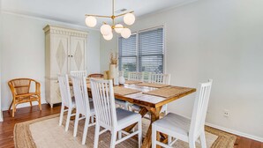 Un comedor con una mesa de madera, sillas blancas, una araña y una ventana con persianas.