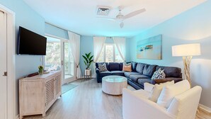 Un salon moderne avec un téléviseur à écran plat, un ventilateur de plafond, un canapé, un fauteuil blanc, une table basse et un tableau accroché au mur.