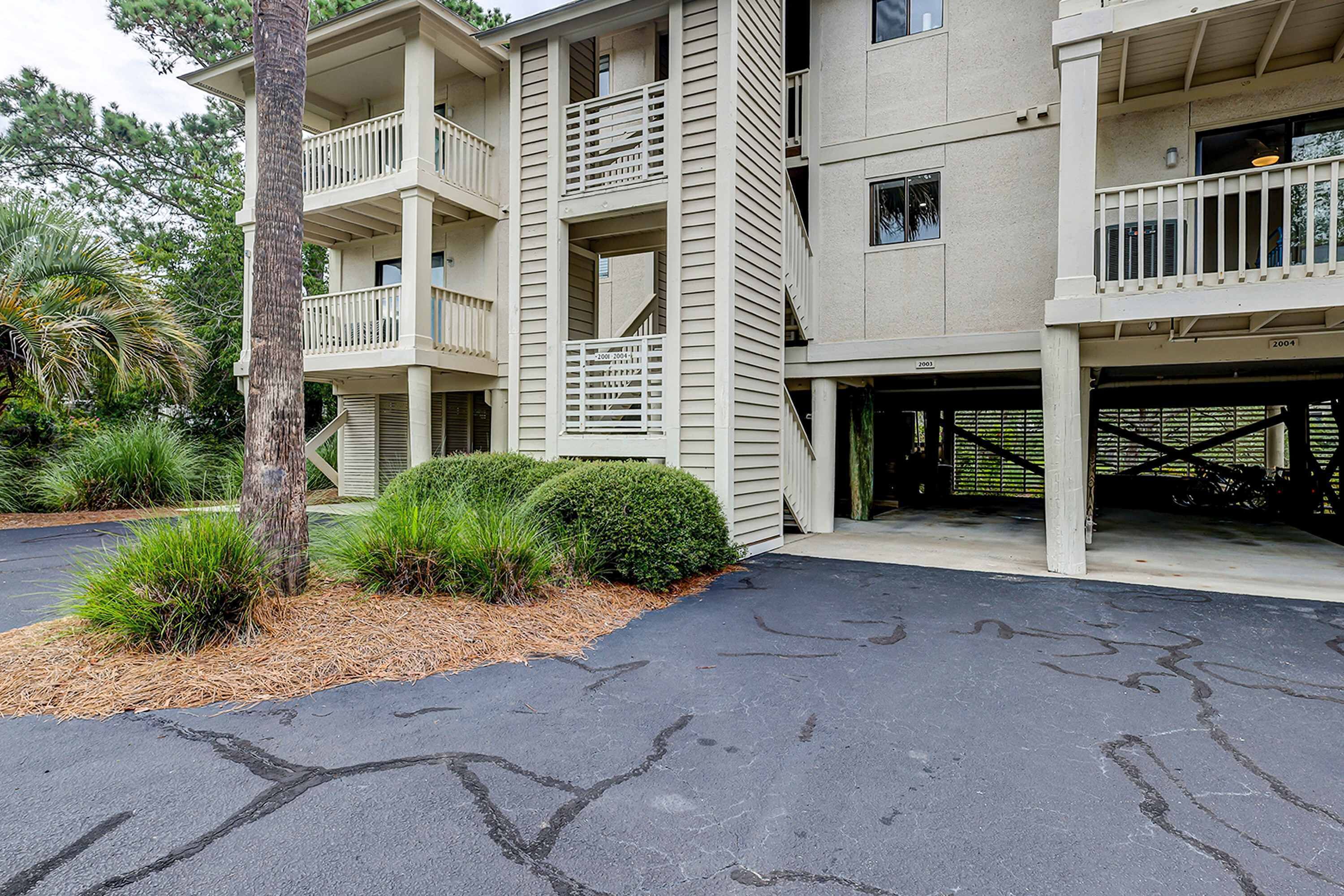 A multi-story building with balconies, a parking area, and surrounding vegetation.