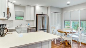 A modern kitchen with white cabinetry, a stainless steel refrigerator, a wooden dining table with white chairs, and a window with blinds. - Scenic view cottage w/ Free Wifi - just a short walk to the beach & park (Kiawah Island)