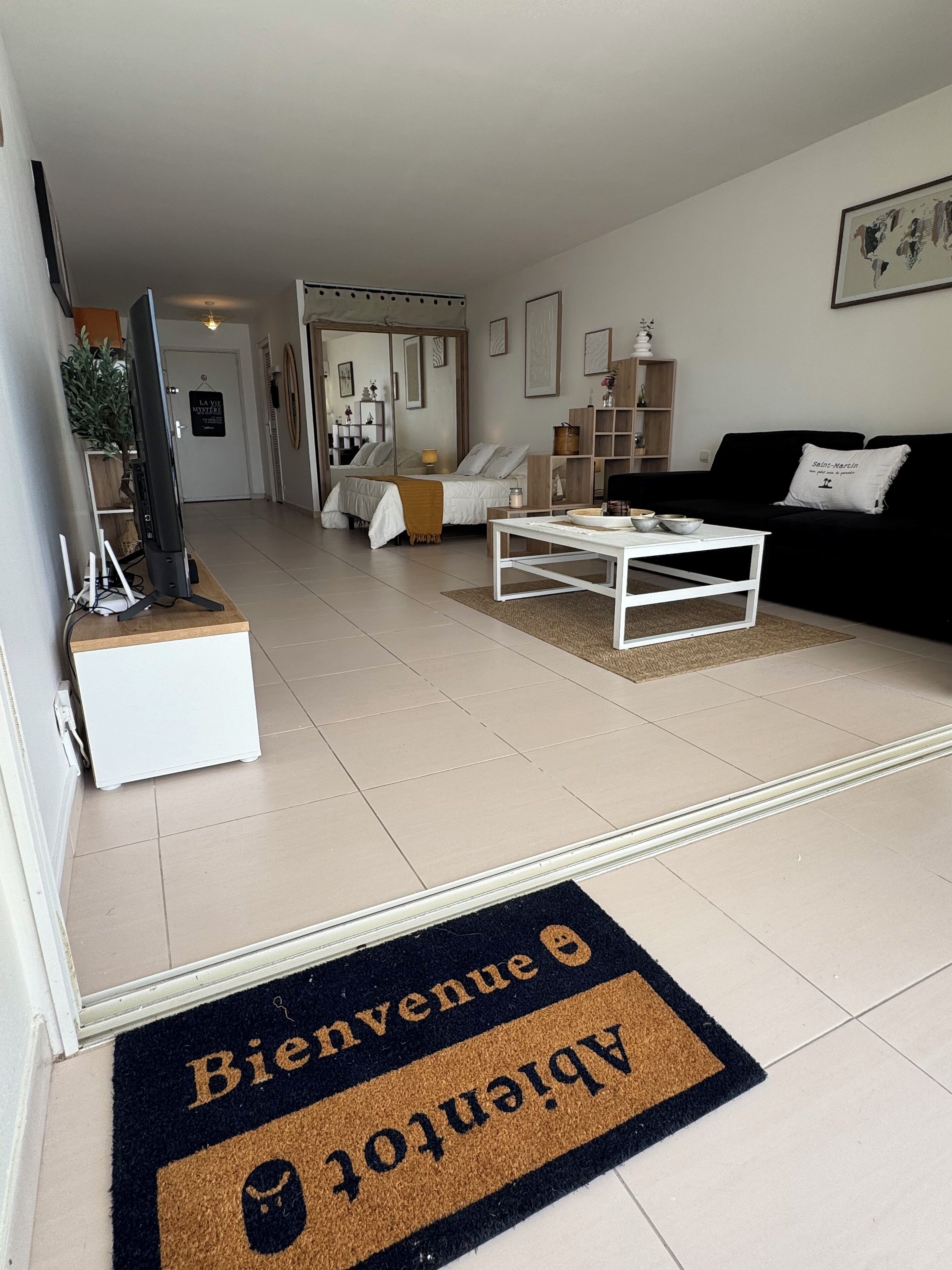 A modern living room with a sofa, coffee table, and a TV on a stand. There is a door with a doormat and a hallway leading to a bedroom.