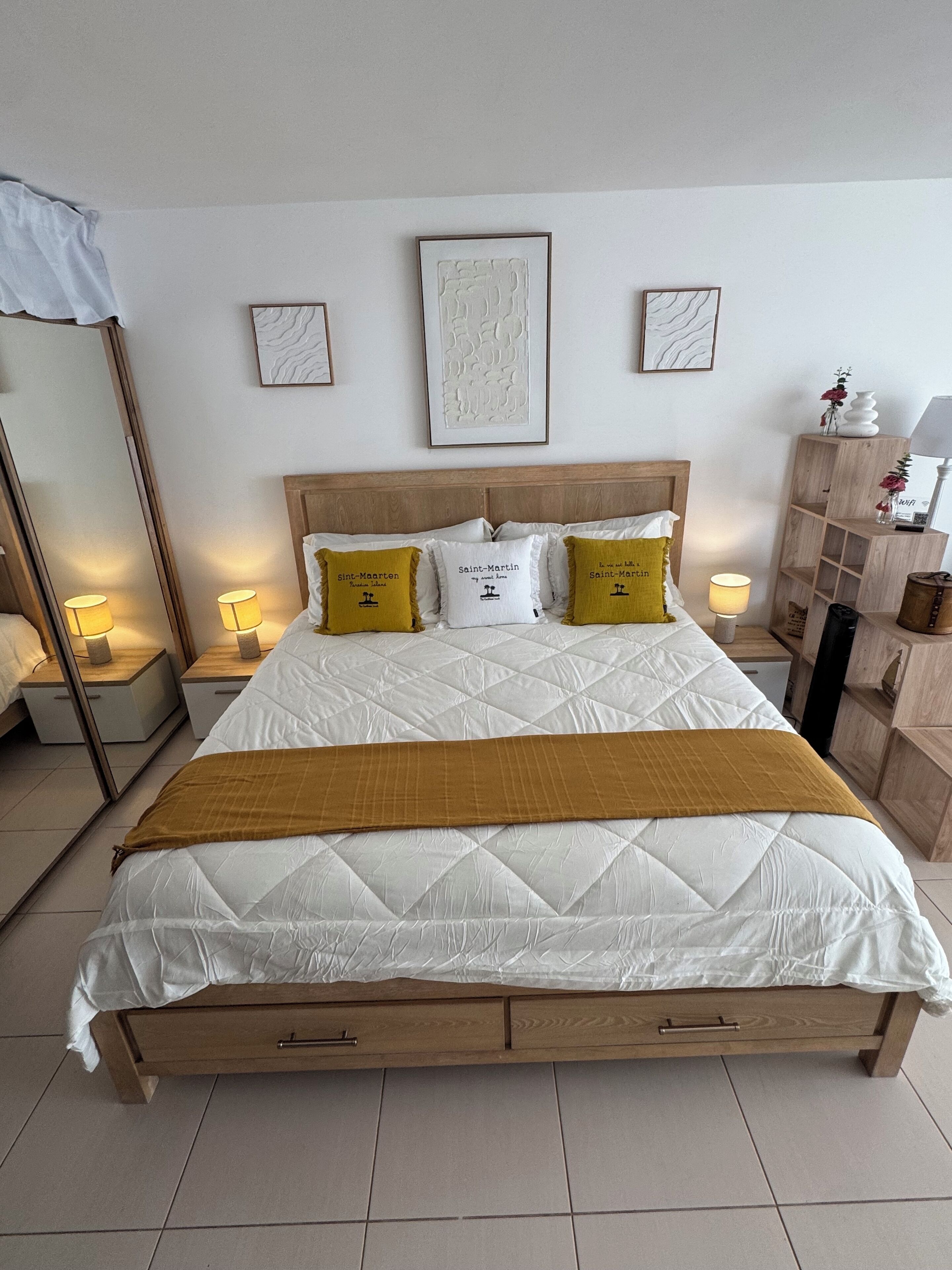 Ein ordentlich bezogenes Bett mit hölzernem Kopfteil, weißer Bettwäsche und zwei senfgelben Kissen. Darüber an der Wand hängen drei gerahmte Kunstwerke.