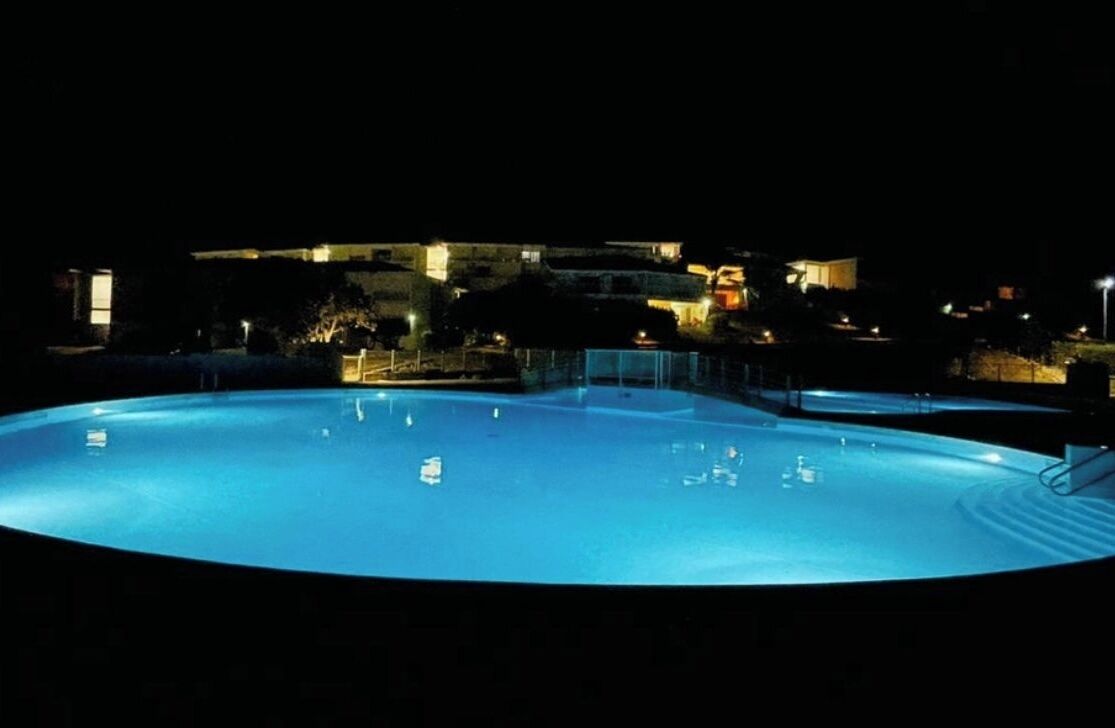 A large, illuminated swimming pool at night with surrounding buildings and a fence.