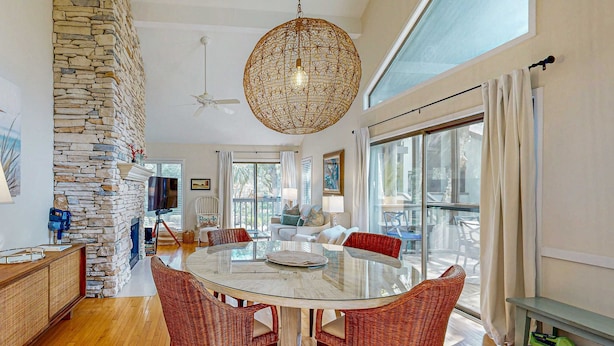 Un vaste espace de vie agrémenté d’une cheminée en pierre, d’une table à manger en verre entourée de chaises en osier et de grandes fenêtres offrant une vue sur l’extérieur.