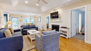 A spacious living room with a ceiling fan, a fireplace, and a TV mounted on the wall. - Dog-friendly home with deck, grill & pool - easy walk to beach, dining & shops (Kiawah Island)