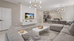Una sala de estar moderna con sofá, mesa de centro y televisor, conectada a una cocina con comedor.