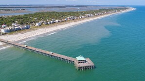Vista aérea de una zona costera con un muelle largo que se adentra en el agua, una playa con casas y un cielo despejado.