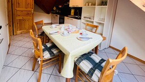 A dining area with a table set for four, wooden chairs with cushions, a wooden door, and a kitchen area in the background.