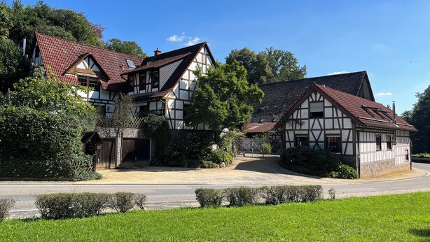 A traditional half-timbered house with a red-tiled roof, surrounded by lush greenery and a clear blue sky.