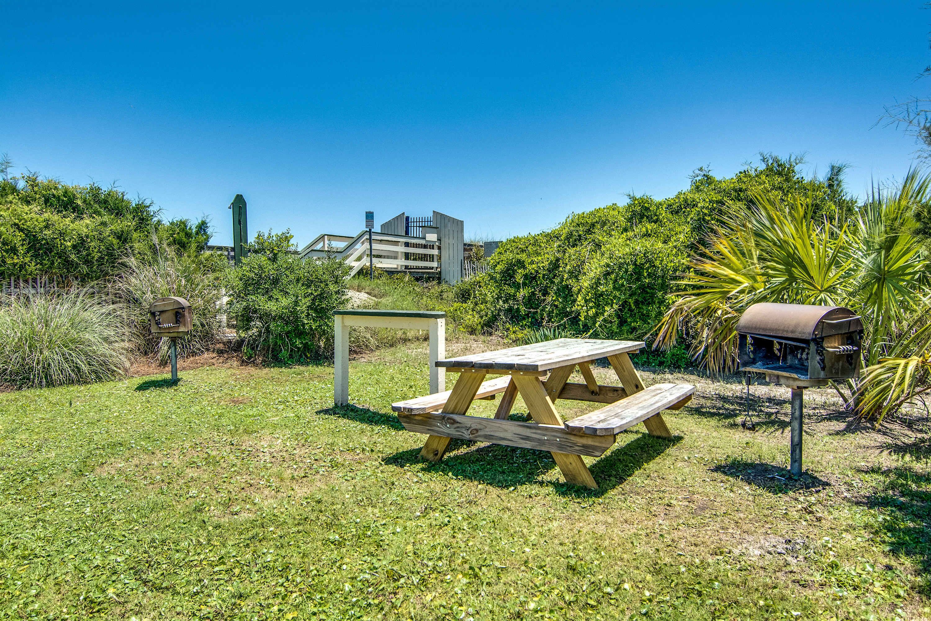 A picnic setup with a wooden table and benches, surrounded by greenery and a barbecue grill.