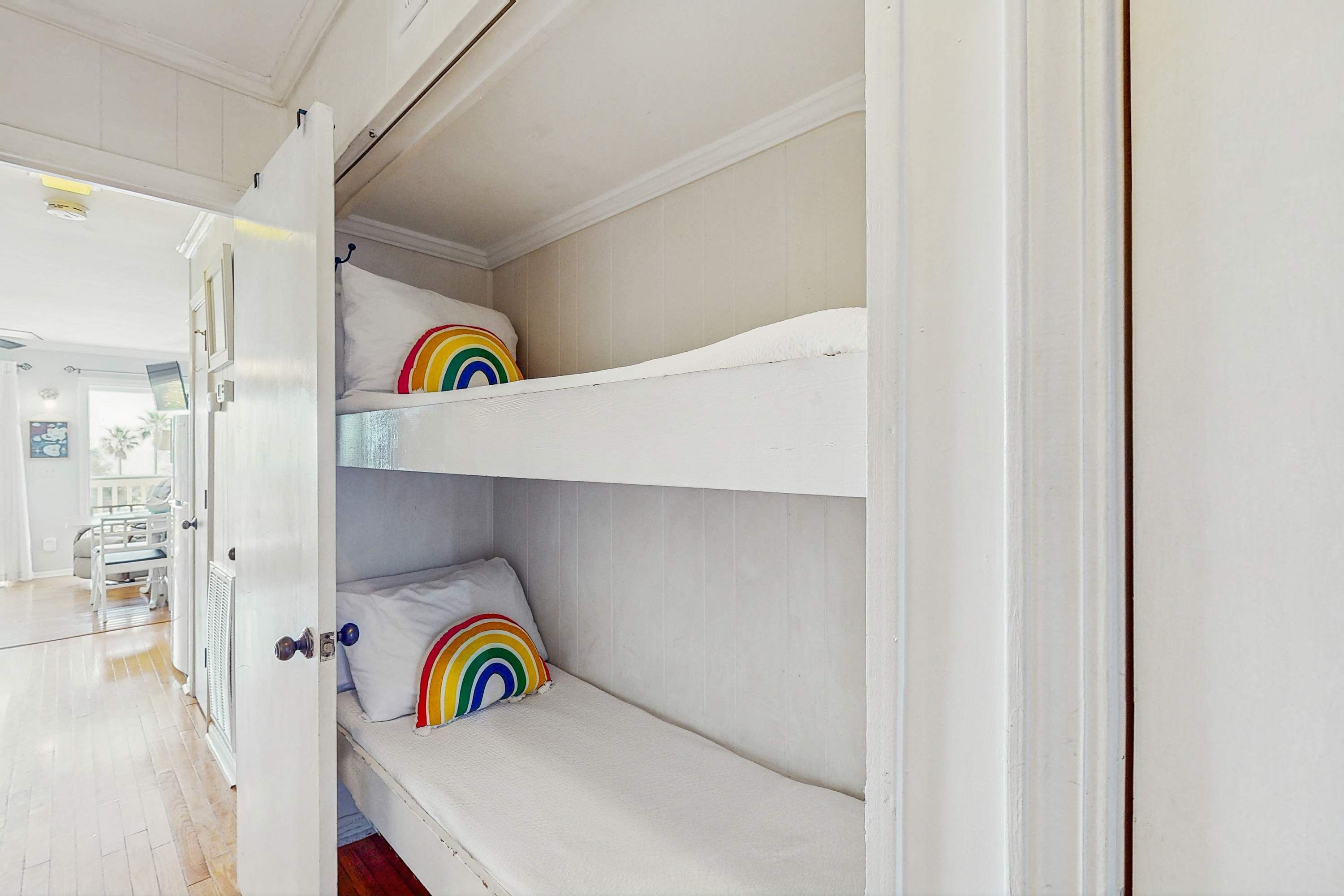 A bunk bed with white bedding and rainbow pillows in a room with wooden flooring and white walls.