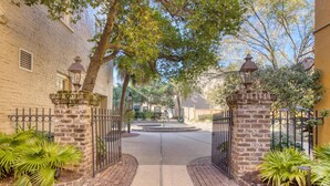 Un camino de entrada con puerta de acceso que conduce a una zona residencial con muros de ladrillo, un sendero central y una pequeña fuente.