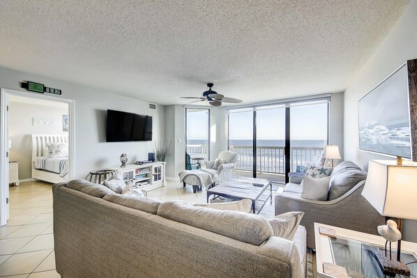 A spacious living room with a large gray sofa, a coffee table, and a ceiling fan. There is a television mounted on the wall and a large window providing a view of the outdoors. - Oceanfront villa with incredible views, balcony & 2 community pools (Folly Beach)