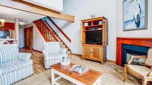 A living room with striped sofas, a wooden coffee table, a television on a wooden cabinet, a fireplace, and a staircase leading to another room. - Three-story ocean-view villa with pool, balcony, charcoal grill, & central AC (Isle of Palms)