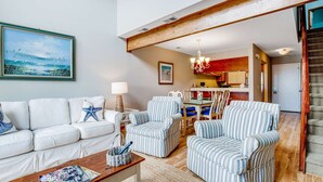 A living room with a white sofa, striped armchairs, a coffee table, and a painting of a beach scene. - Three-story ocean-view villa with pool, balcony, charcoal grill, & central AC (Isle of Palms)