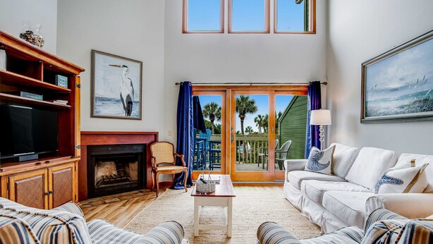 A living room with a fireplace, a television, a sofa, and a view of the outdoors through a sliding glass door. - Three-story ocean-view villa with pool, balcony, charcoal grill, & central AC (Isle of Palms)
