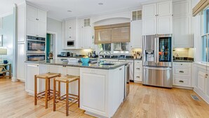 Una cocina amplia con armarios blancos, una isla grande con taburetes, electrodomésticos de acero inoxidable y vistas al exterior.