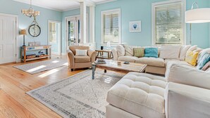 Un salón espacioso con un sofá grande color beige, una mesa de centro y una alfombra estampada. Hay dos sillones y una pequeña mesita de noche. Las paredes son de color azul claro y el suelo es de madera.