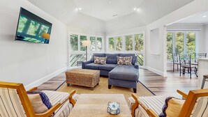 A spacious living room with a sectional sofa, a flat-screen TV mounted on the wall, and large windows offering a view of the outdoors. - Cozy Kiawah Island villa centrally located near local attractions (Kiawah Island)