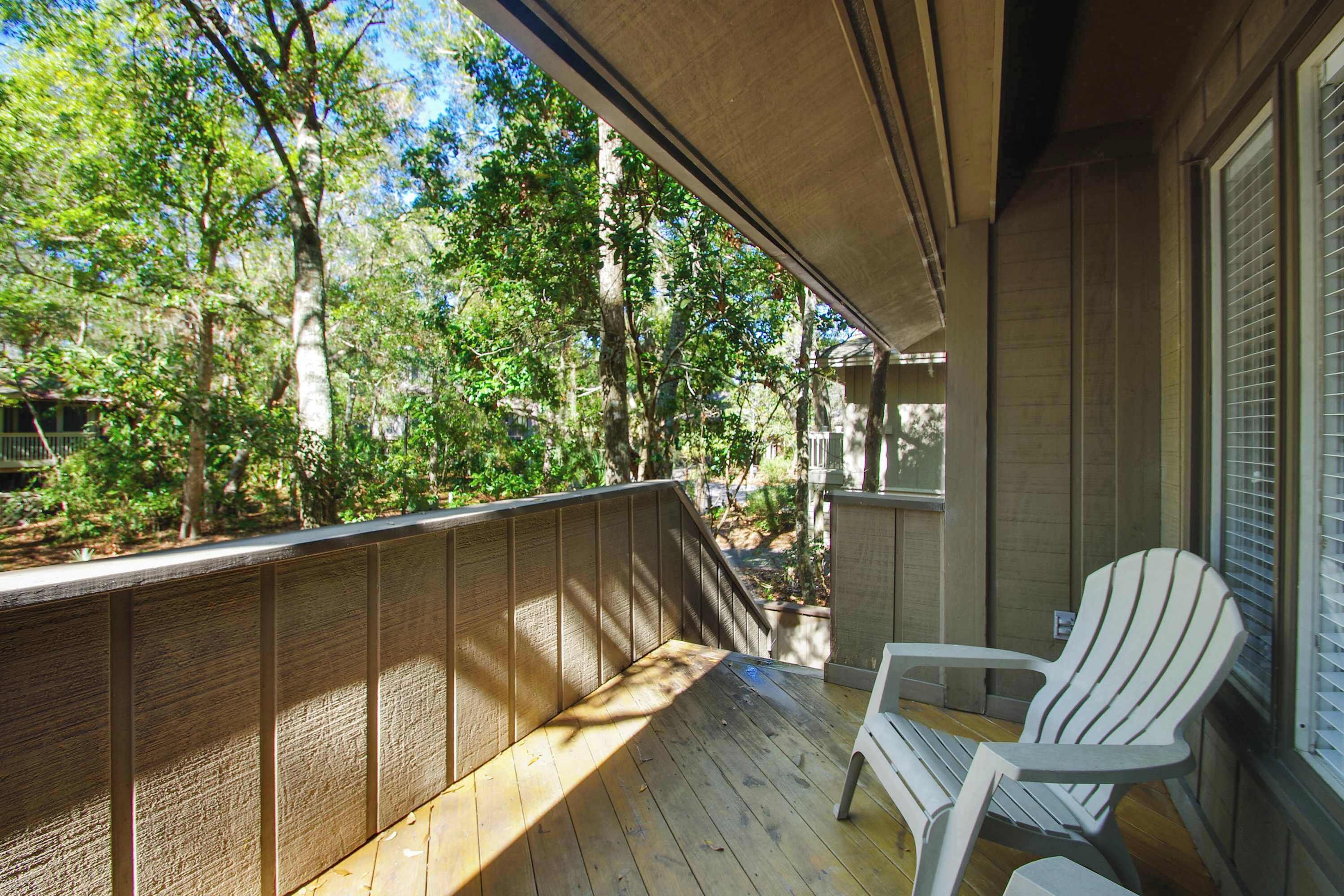 A wooden deck with a railing, a white Adirondack chair, and a view of trees and a house in the background.